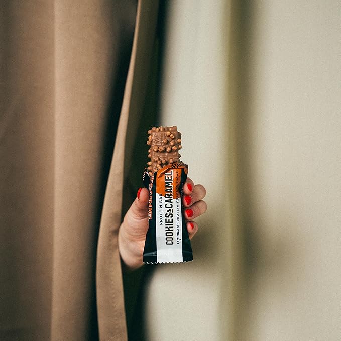 Hand holding a cookies and caramel flavored snack bar against a neutral background Muscle Mecca