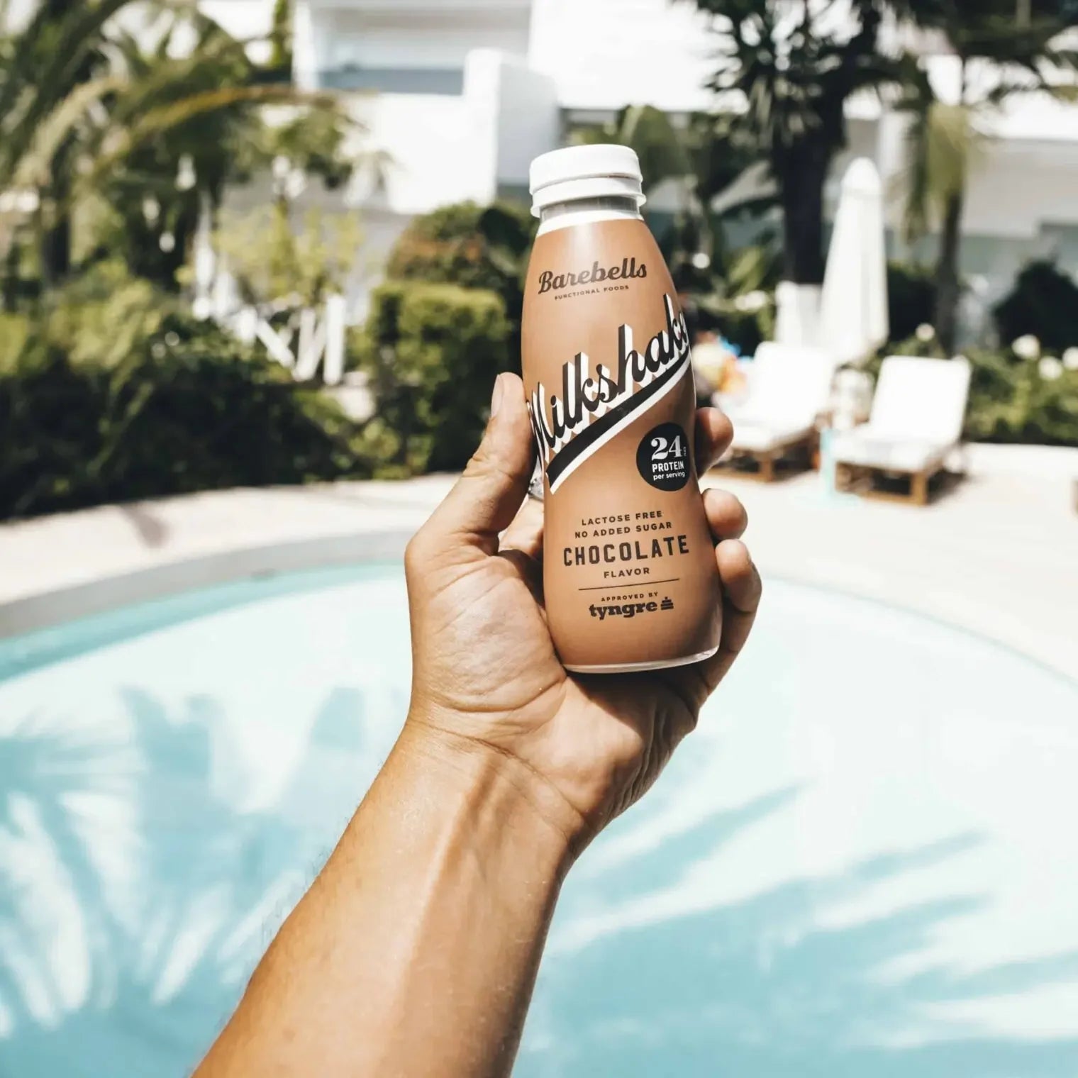 Hand holding a Barebells Milkshake chocolate protein shake bottle by a pool Muscle Mecca