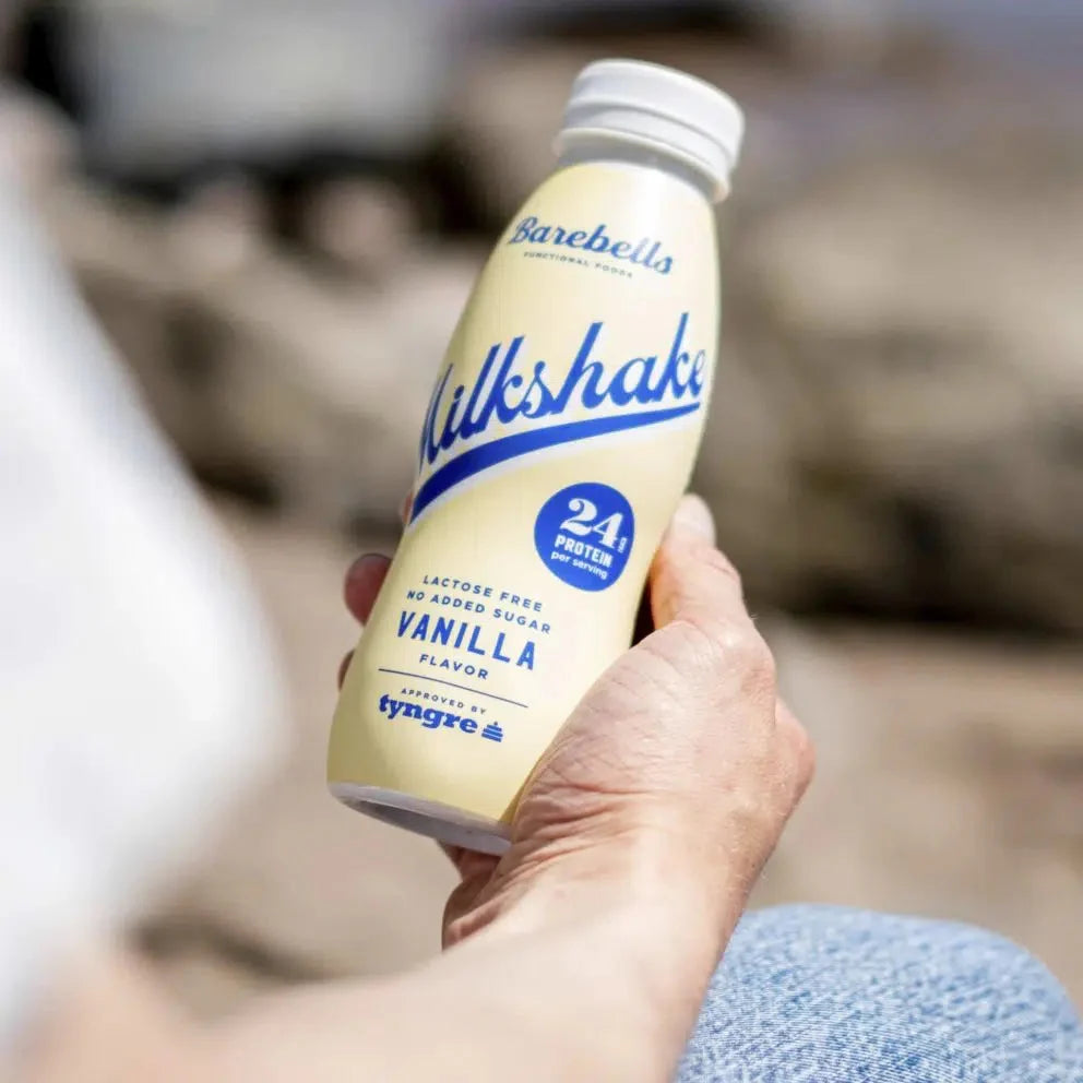 Hand holding a vanilla milkshake bottle with 'Barbella' branding against a blurred natural background. Muscle Mecca