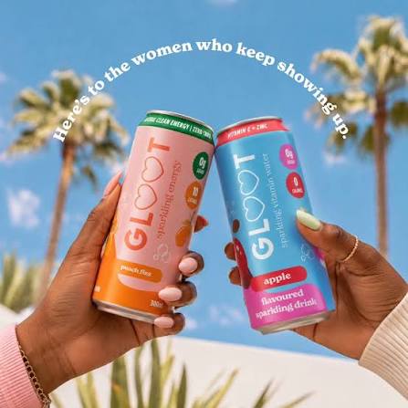 Two hands holding colorful GLOOT sparkling drink cans against a sunny outdoor background with palm trees and a blue sky. White text at the top reads, 'Here's to the women who keep showing up.