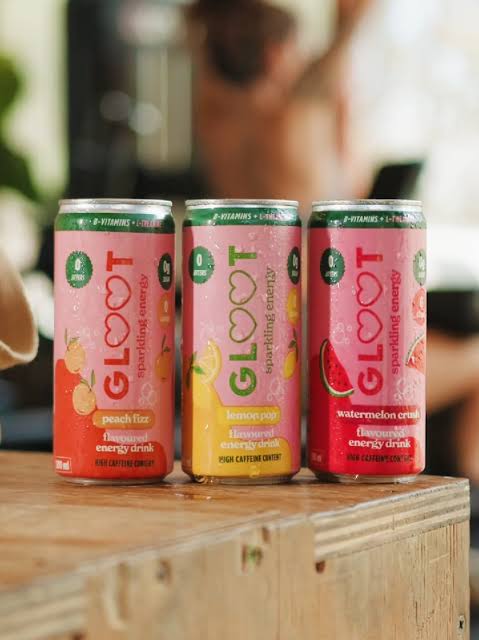 Three GLOOT sparkling energy drink cans in peach fizz, lemon crisp, and watermelon crisp flavors, lined up on a wooden surface with a blurred person in the background.